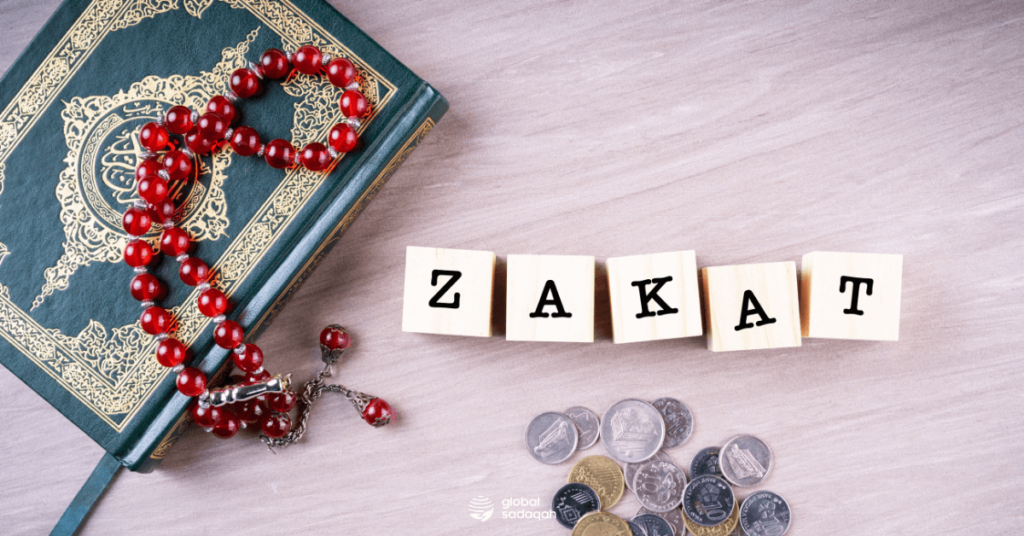  An image of the Quran with a rosary and some coins next to it with the word Zakat in wooden blocks.